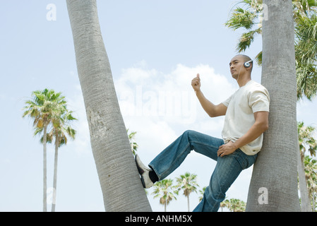 Mann trägt Kopfhörer Baum gelehnt, mit Daumen angehoben, wegschauen Stockfoto