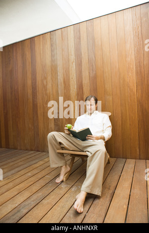 Mann sitzt im Lounge-Stuhl-Lesebuch, Apfel in der hand Stockfoto