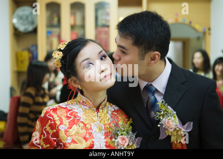 Braut gekleidet in traditioneller chinesischer Kleidung, Blick in die Kamera, als ihr Bräutigam auf die Wange küsst Stockfoto