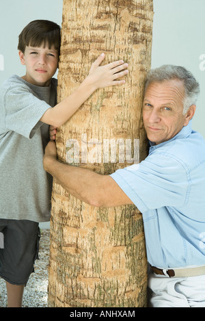 Großvater und Enkel umarmt Baumstamm, beide Blick in die Kamera Stockfoto