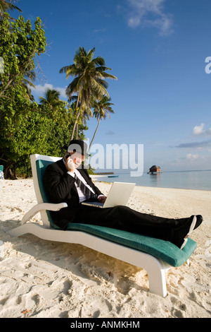 Geschäftsmann in Bowler Hut und Business Anzug auf Laptop-Computer arbeiten, während auf dem Handy am tropischen Strand sprechen Stockfoto