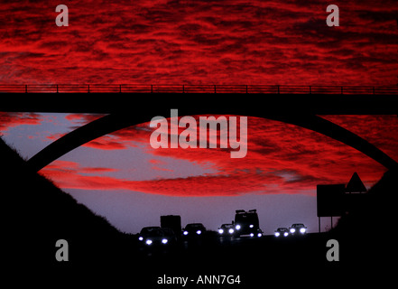 Pendler reisen bei Sonnenuntergang unter einer Autobahnbrücke vor einem strahlend roten Himmel. Bild von Jim Holden. Stockfoto