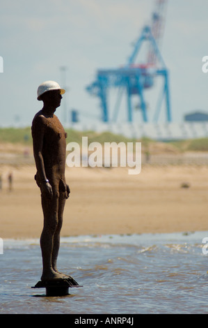 Gusseiserner Mann mit Baumütze, Teil von Antony Gormley's einem anderen Ort am Crosby Beach, der mit einem Ladekran dahinter gesehen wird. Stockfoto