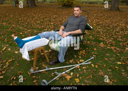 Junger Mann mit einem gebrochenen Bein in Gips, die im Freien auf einem Herbstnachmittag mit abgefallenen Blättern um ihn herum sitzen Stockfoto