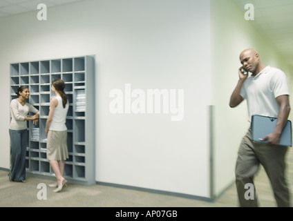 Büro-Szene, zwei Frauen reden von Postfächern während Mann mit Handy-Spaziergänge durch den Flur Stockfoto