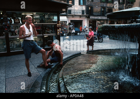 Zwei junge Männer im Westen neben einem Brunnen in Verona Italien einer ständigen und Essen ein Pizzastück, die anderen sitzen Stockfoto