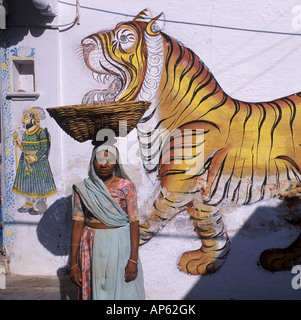 Frau mit Korb auf Kopf vor Rajput Tiger an Wand, Udaipur, Rajasthan, Indien Stockfoto