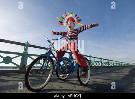 Ein kleiner Junge in einem Indianer Kopfschmuck schreien, wie er auf einem Trike auf den Fahrradweg auf Brighton Seafront spielt Stockfoto