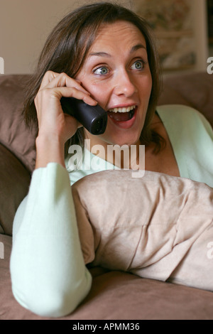Während eines Telefongesprächs während des Gesprächs zu Hause auf dem Sofa entspannen sucht junge attraktive Brünette Frau angenehm überrascht Stockfoto