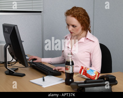 Junge Frau, die arbeitet auf Computer in ihrem Büro-Schreibtisch-Flasche Diät-Koks und Tasche von lo Fett Chips Stockfoto