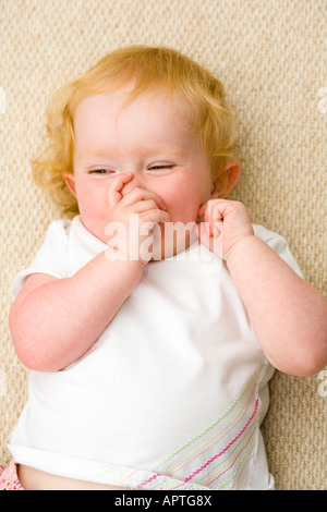 Weibliche Strawberry blonde Kleinkind auf beigen Teppich Daumenlutschen und Lächeln auf den Lippen liegen Stockfoto