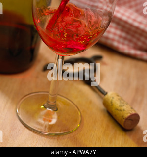 NAHAUFNAHME VON ROTEN ROSEWEIN GEGOSSEN IN GLAS NEBEN KORKENZIEHER UND WEIN FLASCHE MIT ROTEN TISCHDECKEN IM HINTERGRUND Stockfoto