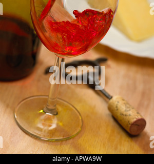 NAHAUFNAHME VON ROTEN ROSEWEIN IN GLAS NEBEN KORKENZIEHER UND WEIN FLASCHE MIT PLATTE KÄSE GEGOSSEN IM HINTERGRUND Stockfoto
