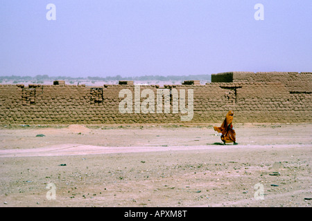 Eine Frau in traditioneller Kleidung zu Fuß neben einem Lehmziegeln Mauern umgebene Dorf am Rande von Agadez Stockfoto