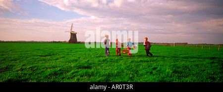 Vier Kinder mit Körben überqueren Ackerland in Holland in der Nähe von Windmühle bei Sonnenuntergang Stockfoto