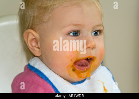 Horizontale Nahaufnahme von einem hübschen kaukasischen Babymädchen sitzen in ihrem Hochstuhl am Abend ihre Lippen lecken Stockfoto