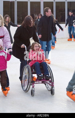 Vertikale Nahaufnahme eines jungen Mädchens in einem Rollstuhl mit viel Spaß um eine Open-Air-Weihnachten-Eisbahn geschoben. Stockfoto