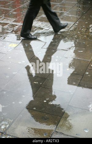 Ein Mann zu Fuß in den Regen halten einen Regenschirm spiegelt sich in der nassen Fahrbahn der Tottenham Court Road, London, UK. Stockfoto