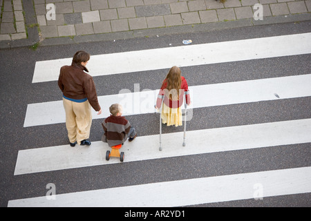 HOLLAND AMSTERDAM ÜBER KOPF DRAUFSICHT DER FRAU MIT ZWEI KINDERN JUNGE AUF TRICICLE MÄDCHEN MIT KRÜCKEN GEHEN ÜBER ZEBRASTREIFEN FÜR FUßGÄNGER Stockfoto