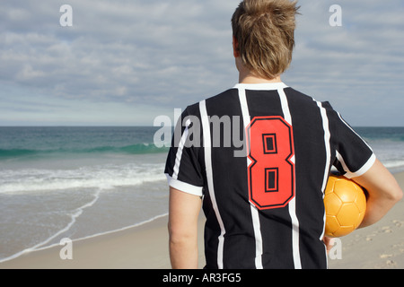 Junger Mann tragen Fußballtrikot halten Fußball Blick auf das Meer Stockfoto