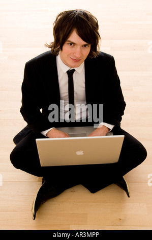 junger Mann auf einem Laptop arbeiten und mit Hilfe eines Mobiltelefons sitzen gekreuzt Beinen auf dem Boden Stockfoto