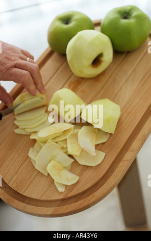 APPLE WIRD IN SCHEIBEN GESCHNITTEN MIT MESSER AUF HÖLZERNEN SCHNEIDEBRETT MIT ÄPFEL UNGESCHÄLT UND GESCHÄLT KOCHEN KOCHEN IM HINTERGRUND Stockfoto