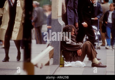 Obdachloser Alkoholiker mit Flasche, der auf dem belebten Einkaufszentrum der Stadtstraße sitzt, auf dem Bürgersteig raucht und trinkt, ignoriert von Passanten Stockfoto