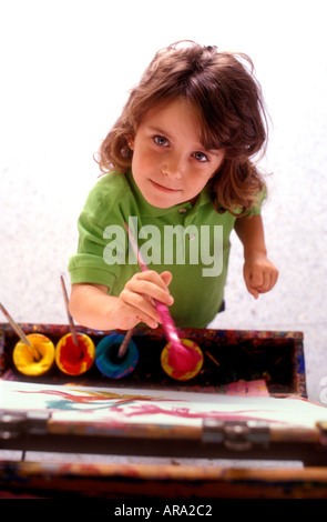 Mädchen 4-6 Jahre mit Farben und Pinsel an der Staffelei in der Schule, die mit einem sanften, selbstbewussten Lächeln aufblickt Stockfoto