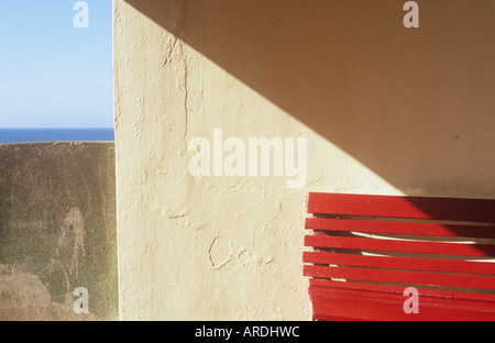 Detail der eine rote Sitzbank in einem cremefarbenen Tierheim mit Diagonale Schatten Betonwand und fernen blauen Meer und Himmel Stockfoto