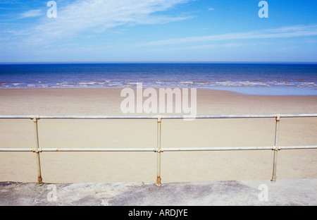 Blick von der Promenade über Geländer zu einem weiten leeren Sandstrand und entfernte ruhige blaue Meer und Himmel Stockfoto