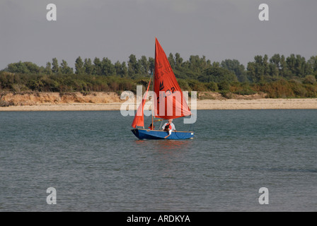 Ein kleines blaues Segelboot mit einem roten Segel kreuzt Eingang zum Hafen von Chichester Stockfoto