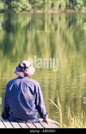 Rückansicht eines Mannes im blauen Hemd mit Sonnenhut sitzen auf Holzsteg am See Stockfoto