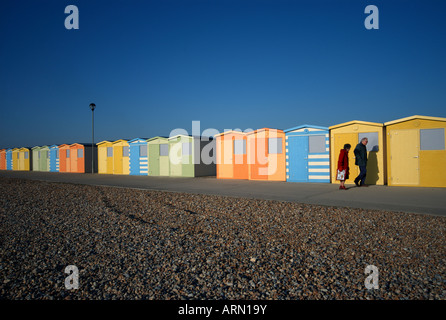 zwei Menschen gehn vorbei an einer Reihe von bunten Strandhäuschen entlang einem Kiesstrand in Seaford, Newhaven, Kent, UK. Stockfoto