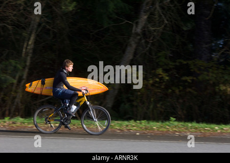 Junger Mann fährt Rad mit Surfbrett in Tofino, Vancouver Island, British Columbia, Kanada. Stockfoto