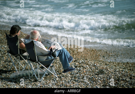 Ein älteres Ehepaar sitzt in Liegestühlen lesen Zeitungen auf einem Kiesstrand am Meer am East Wittering in Sussex Stockfoto