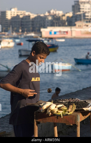 Ägyptischer Mann Maiskolben Grillen über Holzkohle auf dem Bürgersteig mit Fischerbooten im Hintergrund in Alexandria Ägypten Stockfoto