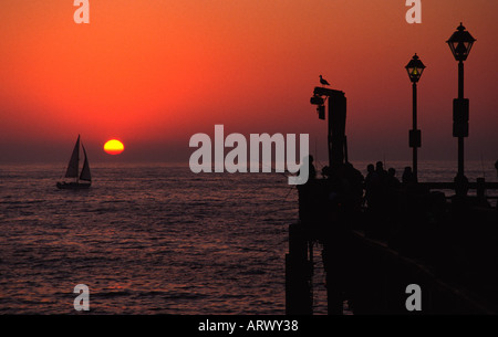 Silhouette eines Fischers auf einem Pier bei Sonnenuntergang mit einem Segelboot in der Ferne Redondo Beach Kalifornien Stockfoto