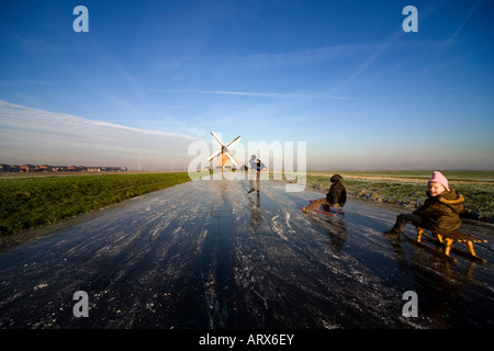 Winter in den Niederlanden. Vater auf Schlittschuhen zieht seine beiden Kinder mit Schlitten oder Schlitten über das Eis in Richtung einer Windmühle. Stockfoto