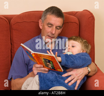 Großvater oder älterer Vater liest vor Sohn Enkel auf seinem Schoß, der müde oder krank sein könnte, Babymilchflasche im Mund Stockfoto