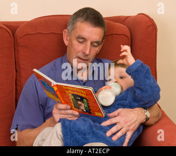 Großvater oder älterer Vater liest vor Sohn Enkel auf seinem Schoß, der müde oder krank sein könnte, Babymilchflasche im Mund Stockfoto