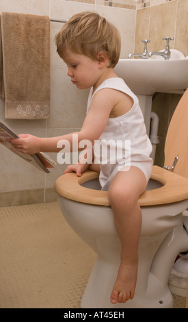 Junge, 2 Jahre alt, sitzt auf Toilette, Toilette, liest Zeitschrift, trägt eine weiße Einzelweste Stockfoto