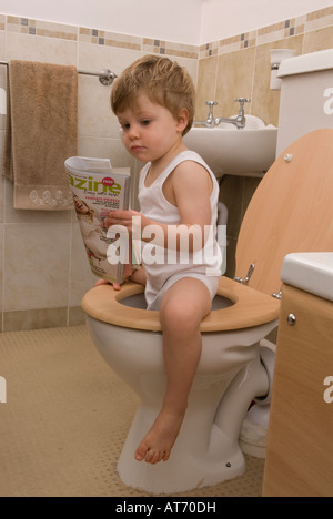 Junge, 2 Jahre alt, sitzt auf Toilette, Toilette, liest Zeitschrift, trägt eine weiße Einzelweste Stockfoto