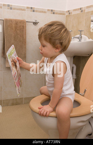 Junge, 2 Jahre alt, sitzt auf Toilette, Toilette, liest Zeitschrift, trägt eine weiße Einzelweste Stockfoto