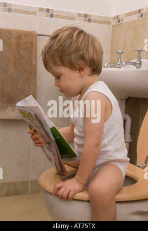 Junge, 2 Jahre alt, sitzt auf Toilette, Toilette, liest Zeitschrift, trägt eine weiße Einzelweste Stockfoto