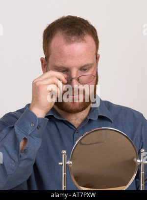 Junger Mann, der eine Brille trägt, in einen kleinen Spiegel schaut, eine Brille für ihn, eine Brille im Spiegel, eine Eitelkeit Stockfoto