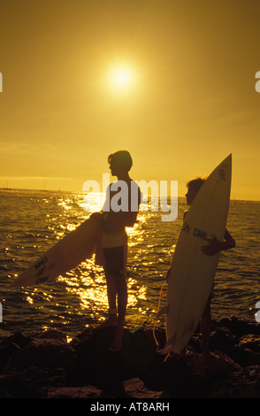 Zwei Surfer mit ihren Brettern stehend auf Lavafelsen Silhouette gegen eine goldene Abendsonne. Stockfoto