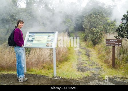 Eine Frau steht neben einem Schild an einem Ausgangspunkt in Hawaiʻi-Volcanoes-Nationalpark. Vulkanische Dampf steigt im Hintergrund. Stockfoto