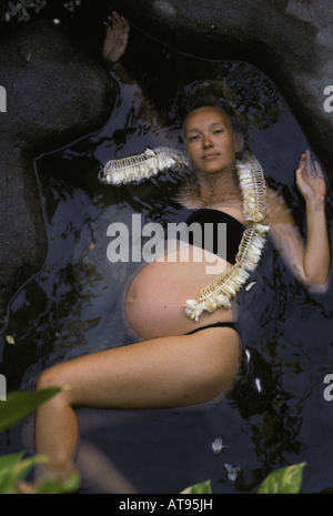 Schwangere Frau schweben im Süßwasser-Pool umgeben von schwarzen Felsen tragen Ingwer Blume Lei. Stockfoto