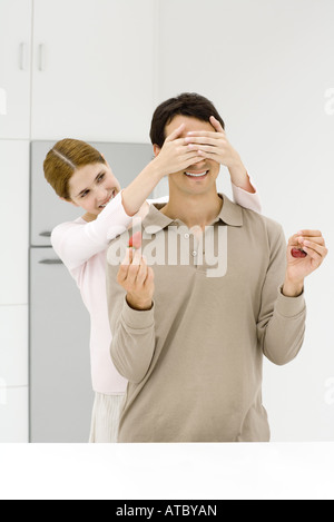 Frau stehend hinter Mann mit ihren Händen über die Augen, Mann, der Erdbeere in der Hand, Kirschen in der anderen hält Stockfoto