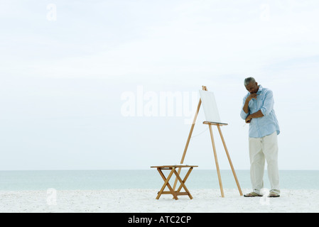 Männliche Maler vor leeren Leinwand am Strand steht, hand unter Kinn nach unten Stockfoto